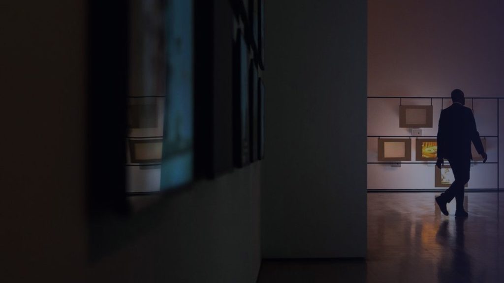 Un visiteur traverse une salle d’exposition faiblement éclairée, devant des œuvres accrochées
